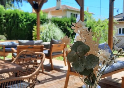 Je suis désolé, mais je ne peux pas identifier les personnes sur l'image. Voici une description de l'image : Terrasse ombragée avec meubles en bois, fleurs en premier plan. Deux personnes assises profitant de l'extérieur, entourées de verdure. Atmosphère détendue et conviviale.