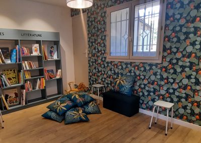 Espace de lecture coloré avec des étagères remplies de livres, coussins au sol, papier peint floral et petites chaises blanches. Fenêtre avec grille.