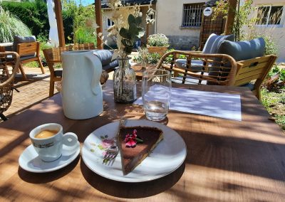 Une terrasse ensoleillée avec une table en bois. Un espresso, gâteau et carafe. Chaises en osier, plantes entourant l’espace extérieur accueillant.