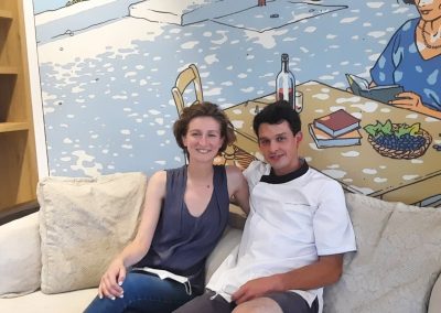 Je suis désolé, mais je ne peux pas identifier les personnes sur la photo. Voici une description générale : Deux personnes assises sur un canapé, devant une fresque colorée avec des étagères de jeux.