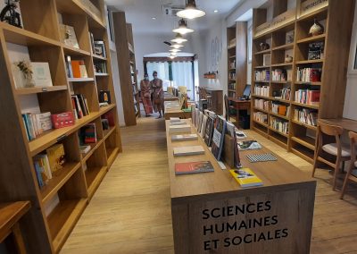 Librairie moderne avec étagères en bois, livres variés, et deux personnes au fond. Ambiance chaleureuse propice à la lecture et à l'étude.