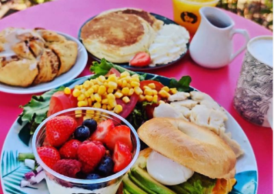 Petit déjeuner coloré sur une table rose : salade, pancakes, pain, fruits, yaourt et boisson dans un jardin lumineux.