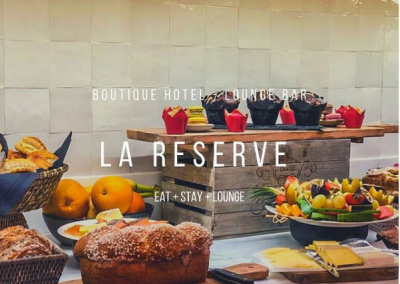 Un buffet coloré avec pâtisseries, fruits et pain dans un hôtel-boutique, étiqueté "La Réserve". Atmosphère accueillante et gourmande présente.