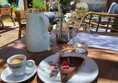Une terrasse ensoleillée avec une table garnie de café, tarte au chocolat, carafe d'eau et vase fleuri, entourée de mobilier confortable.