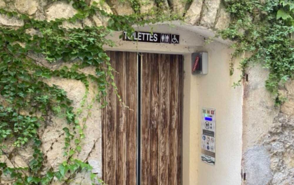 Entrance to wooden public toilets, surrounded by ivy-covered stone walls. The sign above indicates the facilities. No one visible.  