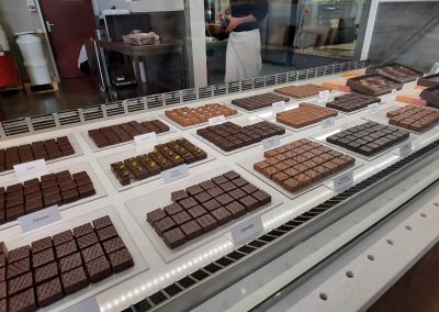 Une vitrine présente des chocolats variés. Un person est en arrière-plan, dans une boutique moderne. Ambiance gourmande et raffinée.