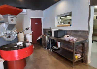 L'intérieur d'une torréfaction avec une grande machine à café rouge et grise. Une personne est visible à travers une fenêtre.