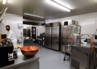 Une personne travaille dans une cuisine industrielle moderne avec divers équipements métalliques, préparant des ingrédients près d'une trancheuse et d'une étagère métallique.