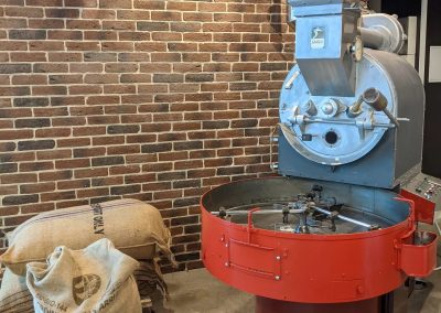 Machine à torréfier rouge avec des sacs de cacao en toile de jute à côté, devant un mur en briques marron.