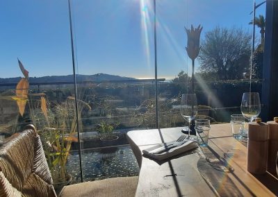 Un restaurant avec vue sur la mer, des verres sur la table, soleil brillant à travers de grandes fenêtres. Ambiance paisible et relaxante.