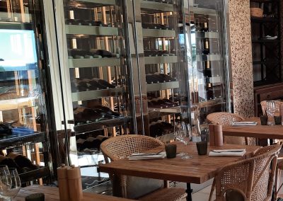 Un restaurant élégant avec des tables en bois, des chaises en osier et des étagères à vin vitrées. Lumière naturelle illuminant l'intérieur calme et accueillant.