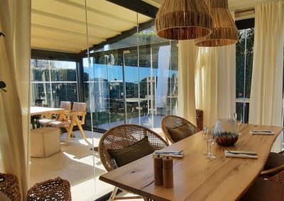 Intérieur moderne d'un restaurant avec des tables en bois, chaises en rotin, grandes fenêtres vitrées, et lumière naturelle abondante. Décoration chaleureuse et conviviale.