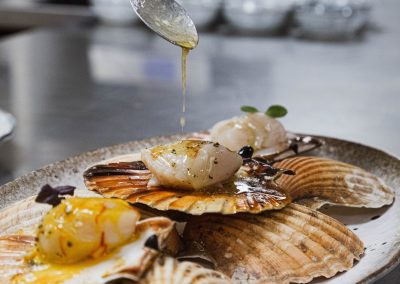 Une personne verse une sauce sur des coquilles Saint-Jacques dans une assiette, située dans une cuisine professionnelle.