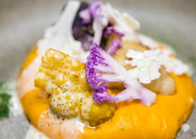 Un plat élégant avec purée orange, choux-fleurs violets et jaunes, nappé d'une sauce blanche onctueuse, dans une assiette grise.