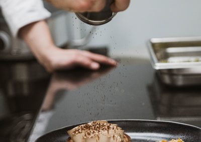 Une personne saupoudre une tarte au chocolat avec du cacao dans une cuisine, sur une assiette noire, avec un plan de travail métallique.