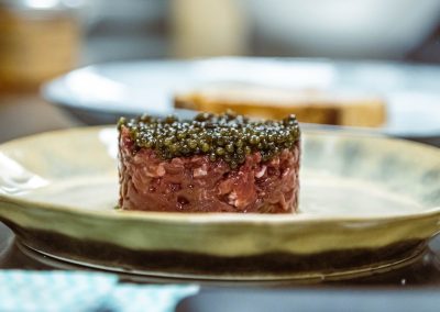 Tartare de bœuf orné de caviar, servi sur une assiette en céramique. En arrière-plan, du pain et un bol flou.