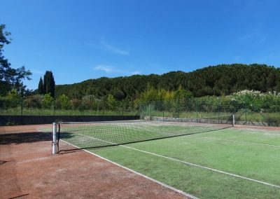 Un court de tennis extérieur entouré de verdure et d'arbres sous un ciel bleu clair, sans personnes présentes sur le terrain.