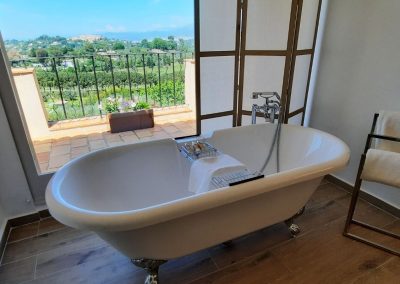 Détail d'une salle de bain avec baignoire, vue sur le jardin et la campagne depuis une terrasse ensoleillée, rideaux en bois à côté.