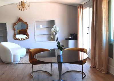 Une pièce élégante avec chaise blanche, miroir doré, table ronde, orchidée, et rideaux beige. Ambiance moderne et lumineuse avec parquet au sol.