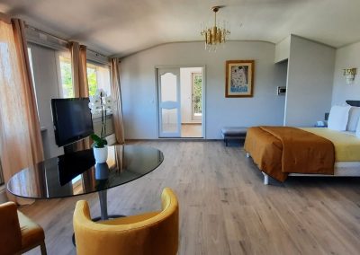 Chambre lumineuse avec lits et bureaux modernes, rideaux beiges, télé, table ronde, chaises jaunes, parquet clair, décoration murale, ambiance confortable et élégante.