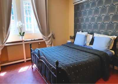 Une chambre avec un lit double, murs décoratifs, rideaux beige, orchidée blanche sur le rebord de la fenêtre, et sol en terre cuite.