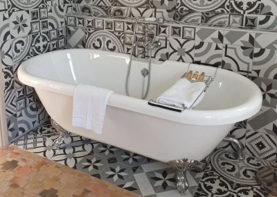 Cette image montre une baignoire blanche classique avec pieds argentés, entourée de carreaux à motifs géométriques noirs et blancs. Serviette blanche en évidence.