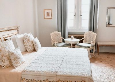 Une chambre lumineuse avec un lit élégant, deux chaises autour d'une table, et un grand miroir. Décor sobre et raffiné.
