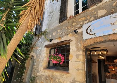 Vieille bâtisse en pierre avec enseigne "La Gourmandise". Fenêtre ornée de fleurs roses. Arbre tropical à gauche. Atmosphère rustique et charmante.