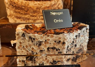 Bloc de nougat oréo exposé dans une vitrine, parsemé d'amandes et de morceaux d'oréo. Étiquette indiquant "Nougat Oréo".