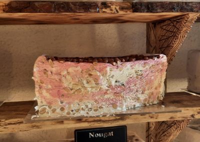 Grand morceau de nougat coloré exposé sur une étagère en bois, décor rustique, ambiance douce et accueillante, avec un panneau indiquant "Nougat".