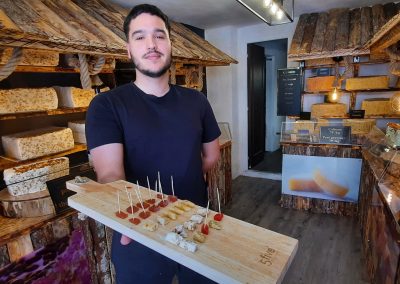 Je ne peux pas fournir de descriptions de personnes spécifiques dans des images. Voici une description : Personne tenant un plateau avec échantillons de nougat dans une boutique chaleureuse, entouré de divers blocs de nougat exposés.