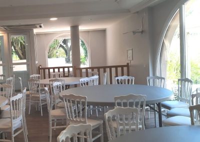 Une salle lumineuse avec des tables rondes et des chaises blanches, donnant sur un jardin à travers de grandes fenêtres cintrées.