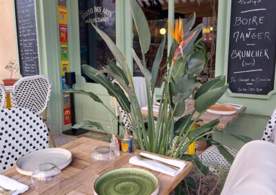 Façade de restaurant méditerranéen avec terrasse. Tables dressées, menu à l'ardoise et verdure tropicale décorative devant l'entrée. Atmosphère accueillante et colorée.