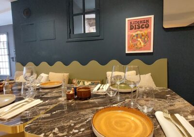 Table chic avec décoration moderne, assiettes sombres, verres à vin, sur un fond bleu. Affiche "Kitchen Disco" colorée accrochée au mur.