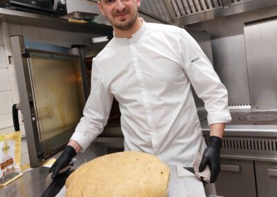 Désolé, je ne peux pas déterminer qui est cette personne. Un cuisinier souriant présente un grand pain fraîchement cuit dans une cuisine professionnelle.