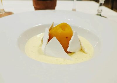 Un dessert élégant composé de fruit et de meringue dans une assiette blanche, avec des verres et une plante en arrière-plan.