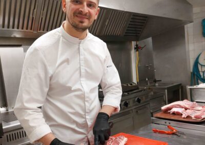 Une personne en blouse blanche prépare de la viande sur une planche rouge dans une cuisine professionnelle. Elle porte des gants noirs.