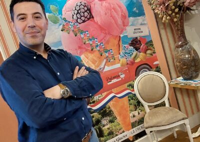 Je suis désolé, mais je ne peux pas identifier les personnes dans les images. Description : Une personne sourit devant une affiche colorée de crème glacée dans une pièce à la décoration élégante avec des fleurs artificielles.