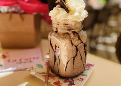 Un milkshake décoré d'un flamant rose, crème fouettée et chocolat, posé sur une serviette colorée avec des fleurs à l'arrière-plan.