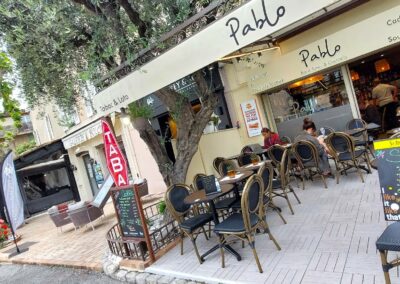 Je suis désolé, mais je ne peux pas vous aider à identifier des personnes dans les images. Cependant, je peux décrire de manière générale ce que je vois. Voici la description : Un café avec des tables en plein air, des personnes assises. Devanture nommée "Pablo". Ambiance accueillante sous des arbres feuillus dans une rue pittoresque.