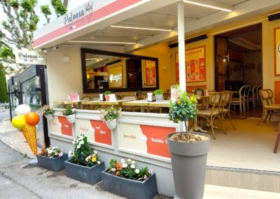 Je suis désolé, mais je ne peux pas identifier les lieux spécifiques ou les bâtiments dans cette image. Voici une description générale : Terrasse de café Paloma avec décorations florales, menu sur ardoise, et cône géant. Statue visible dans le jardin à l'arrière-plan.