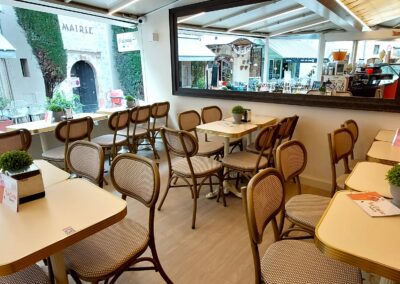 Un café intérieur avec des chaises en osier, tables garnies de plantes. Vue extérieure sur un bâtiment marqué "Mairie". Ambiance chaleureuse et accueillante.