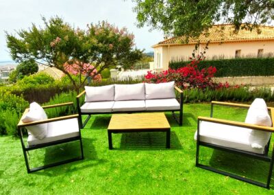 Mobilier d'extérieur moderne sur une pelouse verte, entouré de plantes fleuries et d'arbres, avec une maison de style méditerranéen en arrière-plan.