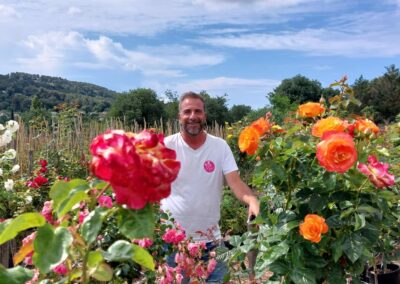 Désolé, je ne sais pas qui est cette personne. Un individu souriant se tient dans un jardin de roses colorées, avec des collines en arrière-plan.