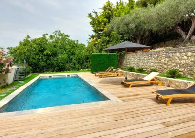 Une piscine entourée de chaises longues en bois, près d'un mur de pierre et d'arbres verdoyants. Ciel dégagé, ambiance paisible.