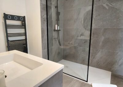 Salle de bain moderne avec douche à l'italienne en verre, carrelage gris, lavabo blanc, mitigeur noir et porte-serviettes chauffant noir. Ambiance épurée et élégante.