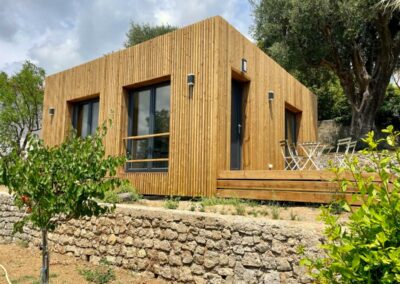 Petite maison en bois moderne entourée de verdure, avec terrasse et table extérieure, sous un ciel nuageux, située dans un cadre paisible.