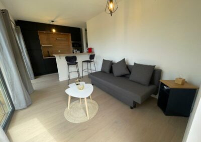 Salon moderne avec canapé gris, table basse blanche, cuisine ouverte avec comptoir, chaises hautes et éclairage suspendu. Ambiance lumineuse et épurée.