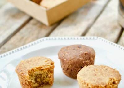 Trois muffins sur une assiette blanche, avec boîte en carton contenant d'autres muffins. Les muffins sont partiellement consommés, sur une table en bois.