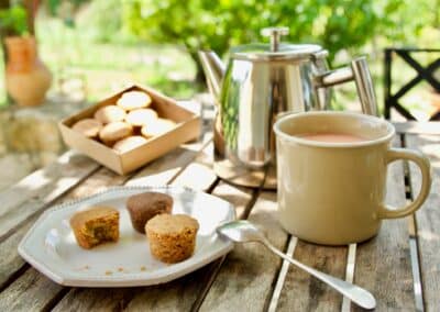 Un plateau de gâteaux, théière en métal, tasse de thé et cuillère sur table en bois, en extérieur, avec fond verdoyant.